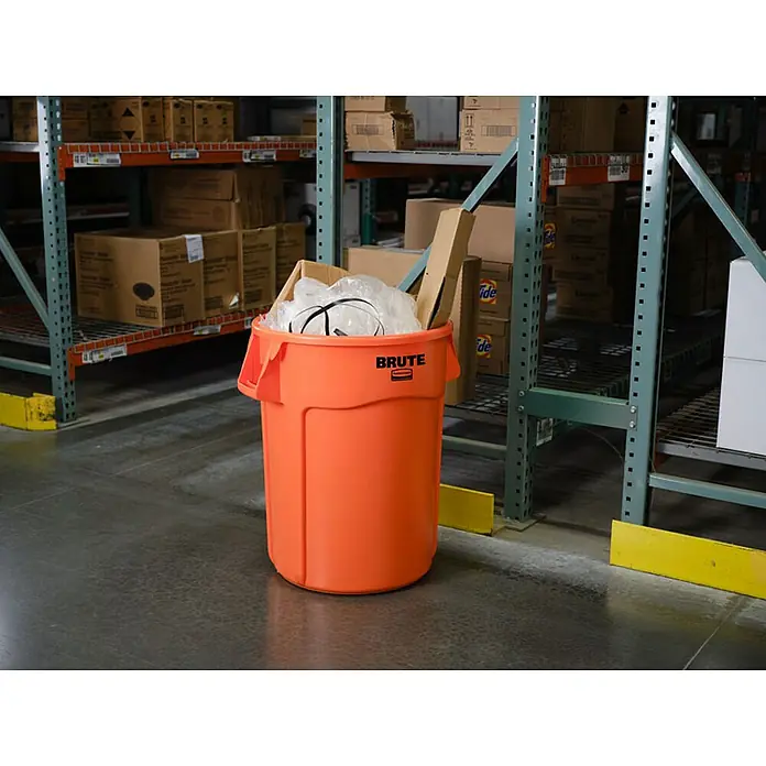 Proregal Standard-Mülleimer Rubbermaid Brute runde Mehrzwecktonne mit Belüftungskanälen - 121,1 l, Kunststoff, Orange - Ein orangefarbener "Brute" Abfallbehälter, voll mit Müll, steht in einem Lagerhaus vor Lagerregalen.