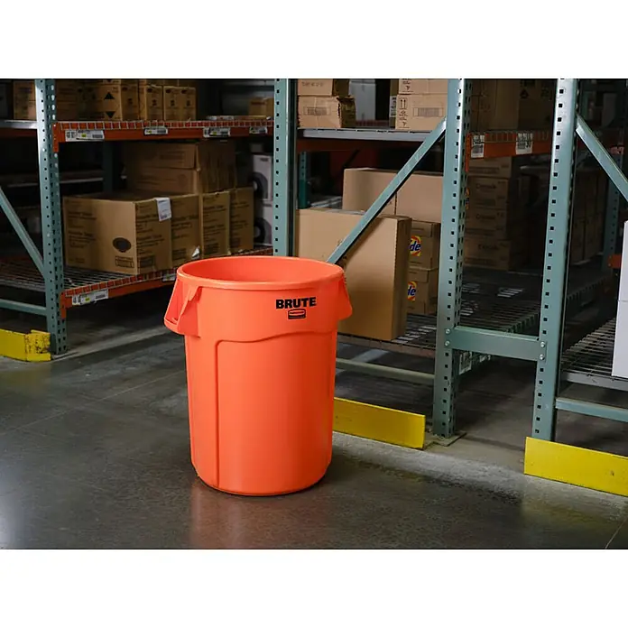 Proregal Standard-Mülleimer Rubbermaid Brute runde Mehrzwecktonne mit Belüftungskanälen - 121,1 l, Kunststoff, Orange - Ein orangefarbener, zylindrischer Brute-Mülleimer aus Kunststoff steht in einem Lagerhaus zwischen den Regalen.