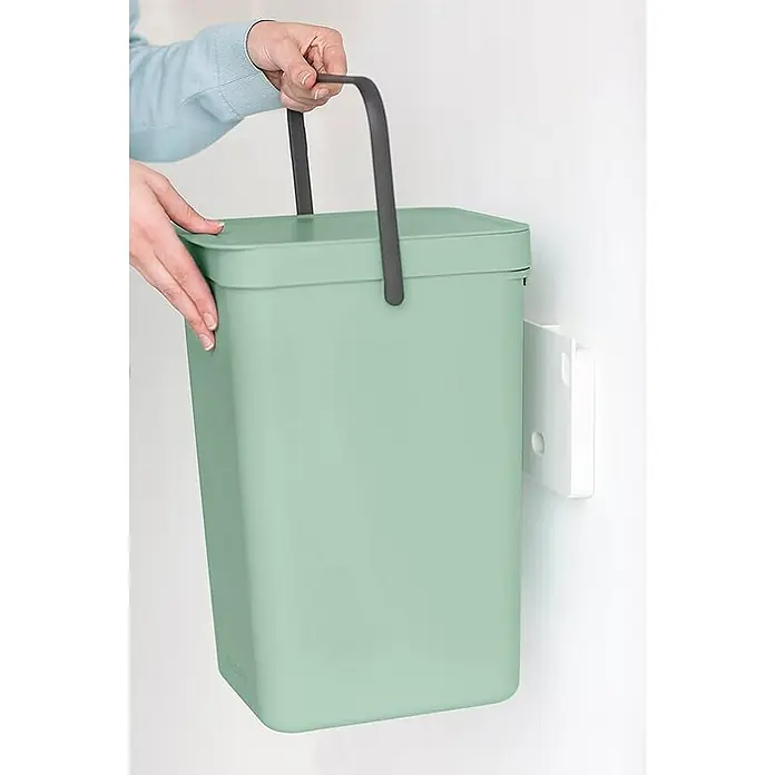 Proregal Standard-Mülleimer Brabantia Abfallsammler mit Stay-open Deckel - 16 l, Kunststoff, Jade - Eine grüne Bio-Mülltonne mit einem grauen Griff wird von einer Person gehalten, während sie an einer Wandhalterung befestigt ist.
