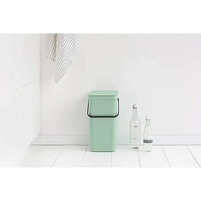 Proregal Standard-Mülleimer Brabantia Abfallsammler mit Stay-open Deckel - 16 l, Kunststoff, Jade - Ein hellgrüner, quadratischer Abfallbehälter mit Deckel steht neben zwei Wasserflaschen vor einer weißen Wand. Ein Küchentuch hängt.
