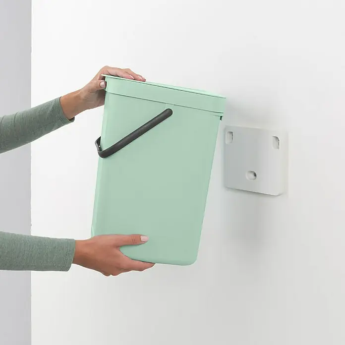 Proregal Standard-Mülleimer Brabantia Abfallsammler mit Stay-open Deckel - 12 l, Kunststoff, Jade - Die Hände einer Person halten einen mintgrünen Bio-Abfalleimer vor einer weißen Wand mit einer weißen Wandhalterung.