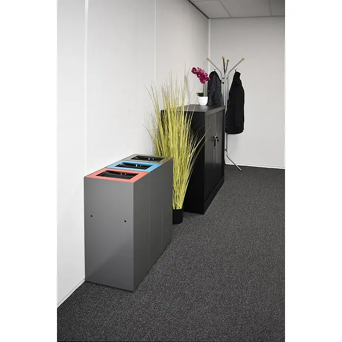 Proregal Mülltrennsystem Robust & modular - 60 l, Stahl, Anthrazit - Grauer Recyclingbehälter mit farbcodierten Öffnungen zur Mülltrennung in einem Büro, daneben ein schwarzer Schrank, Dekoration und eine Garderobe.