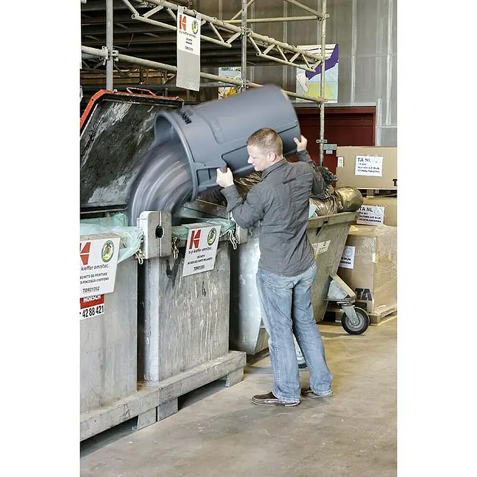 Proregal Standard-Mülleimer Rubbermaid Brute runde Mehrzwecktonne mit Belüftungskanälen - 166,5 l, Kunststoff, Grau - Ein Mann leert einen grauen Müllbehälter in einen großen Abfallcontainer, der mit Firmenlogos versehen ist.