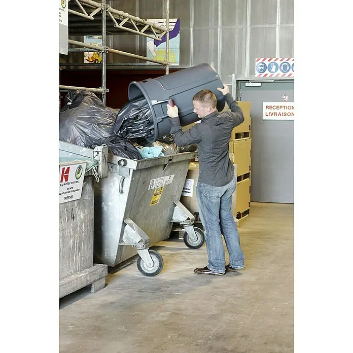 Proregal Standard-Mülleimer Rubbermaid Brute runde Mehrzwecktonne mit Belüftungskanälen - 166,5 l, Kunststoff, Grau - Eine Person leert einen grauen Abfallbehälter in einen großen Müllcontainer, um die Abfallentsorgung und das Recycling zu erleichtern.