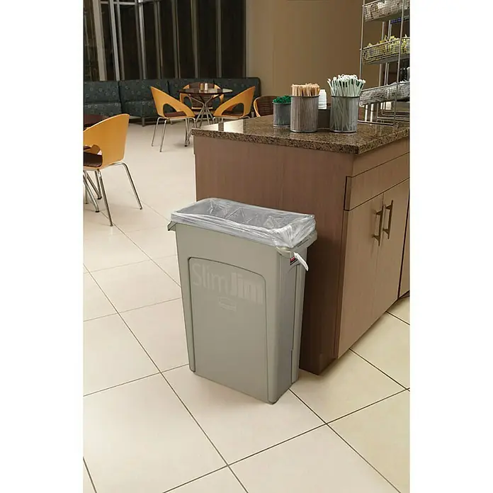 Proregal Standard-Mülleimer Rubbermaid Slim Jim Mülleimer mit Belüftungskanälen - 60 l, Kunststoff, Grau - Grauer Slim Jim-Mülleimer mit durchsichtigem Müllbeutel in einem Wartebereich neben einer braunen Theke.