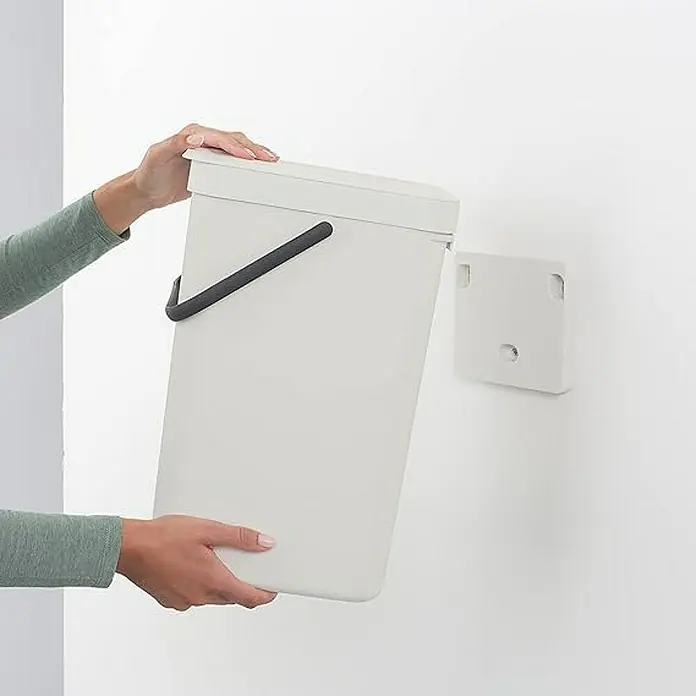 Proregal Standard Mülleimer Brabantia Abfallsammler mit Stay-open Deckel - 16 l, Kunststoff, Hellgrau - Ein cremefarbener Mülleimer mit schwarzem Griff wird in eine passende Wandhalterung eingesetzt. Das Produkt ist für die einfache Müllentsorgung konzipiert.