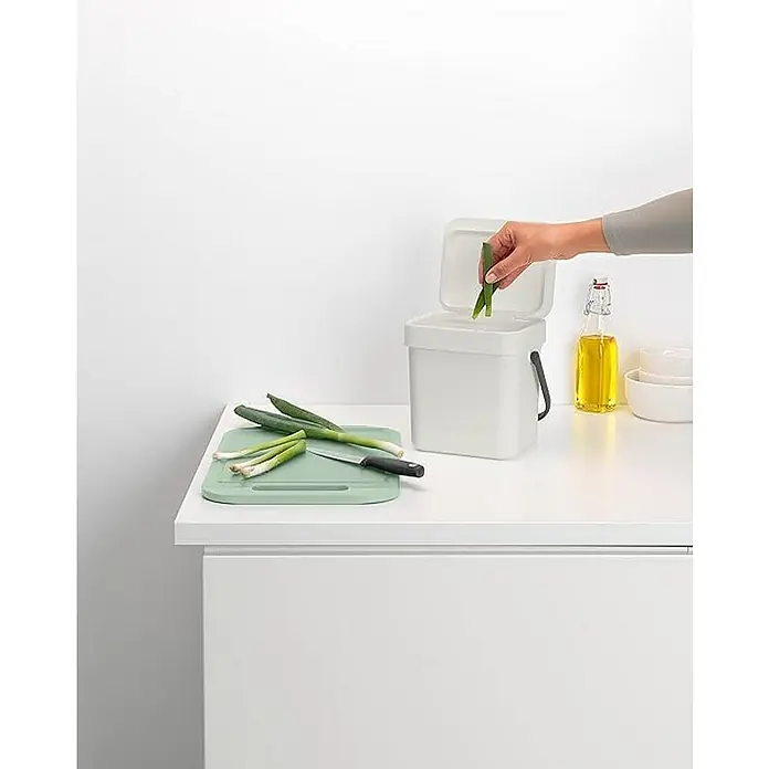 Proregal Standard Mülleimer Brabantia Abfallsammler mit Stay-open Deckel - 3 l, Kunststoff, Hellgrau - Das Bild zeigt einen weißen Bio-Abfalleimer mit einem grünen Schneidebrett, auf dem geschnittene Frühlingszwiebeln liegen. Eine Hand wirft Gemüseabfälle in den Eimer.