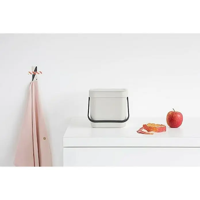 Proregal Standard Mülleimer Brabantia Abfallsammler mit Stay-open Deckel - 3 l, Kunststoff, Hellgrau - Ein minimalistischer, weißer Komposteimer steht auf einer weißen Oberfläche, daneben ein roter Apfel und Apfelschalen. Handtuch hängt an Haken.