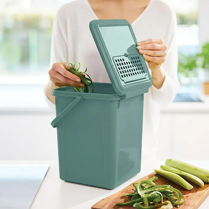 Proregal Standard-Mülleimer Robuster Abfalleimer mit geruchsdichtem Deckel & Kohlefilter - 9 l, Kunststoff, Grün - Eine Frau leert einen grünen Küchenkomposteimer mit Deckel, während sie Gemüsereste von einem Schneidebrett entsorgt.