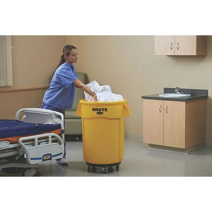 Proregal Standard-Mülleimer Rubbermaid Brute runde Mehrzwecktonne mit Belüftungskanälen - 166,5 l, Kunststoff, Gelb - Eine Krankenschwester leert schmutzige Wäsche in einen gelben Brute-Wäschebehälter mit Rollen in einem Krankenhauszimmer.