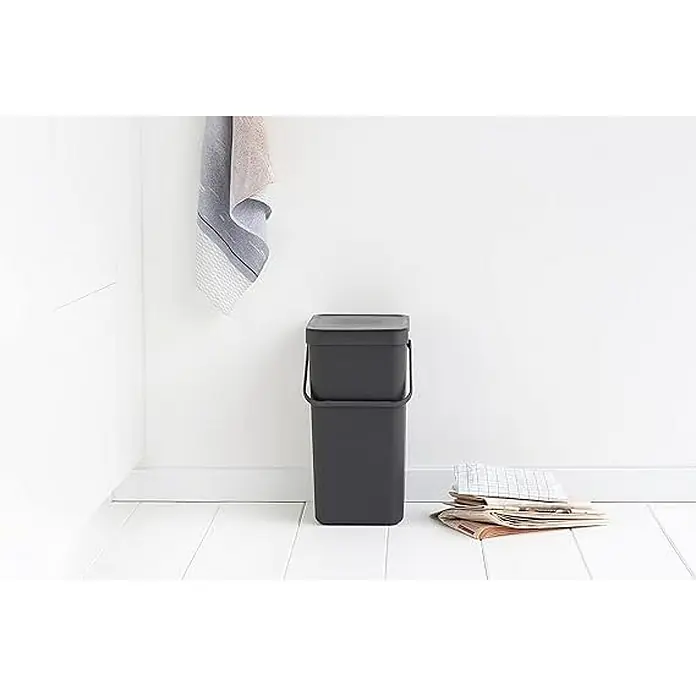 Proregal Standard Mülleimer Brabantia Abfallsammler mit Stay-open Deckel - 16 l, Kunststoff, Dunkelgrau - Ein grauer Brabantia Sort & Go Abfallbehälter mit Deckel steht im Raum, daneben liegt eine Zeitung. Ein Handtuch hängt an der Wand.