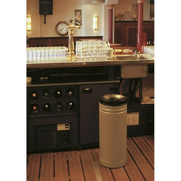 Proregal Papierkorb & Abfallsammler aus Metall selbstlöschend mit doppelter Rauchgasumlenkung - 50 l, Metall, Grau, Kopfteil Schwarz - Ein heller, zylindrischer Mülleimer mit schwarzem Deckel steht vor einer Bar mit Weinkühler, um die Sauberkeit im Barbereich zu gewährleisten.