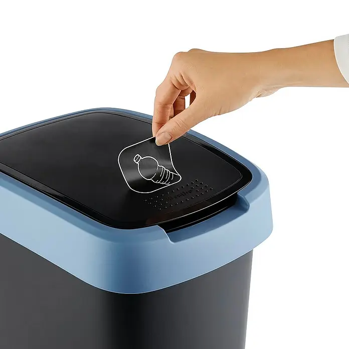 Proregal Standard-Mülleimer Klassischer Abfalleimer für Innenbereich - 25 l, Kunststoff, Schwarz-Blau - Nahaufnahme eines schwarzen EKO-Abfalleimers mit blauem Deckel, auf dem ein Aufkleber mit einer Flasche angebracht ist, der von einer Hand abgezogen wird.