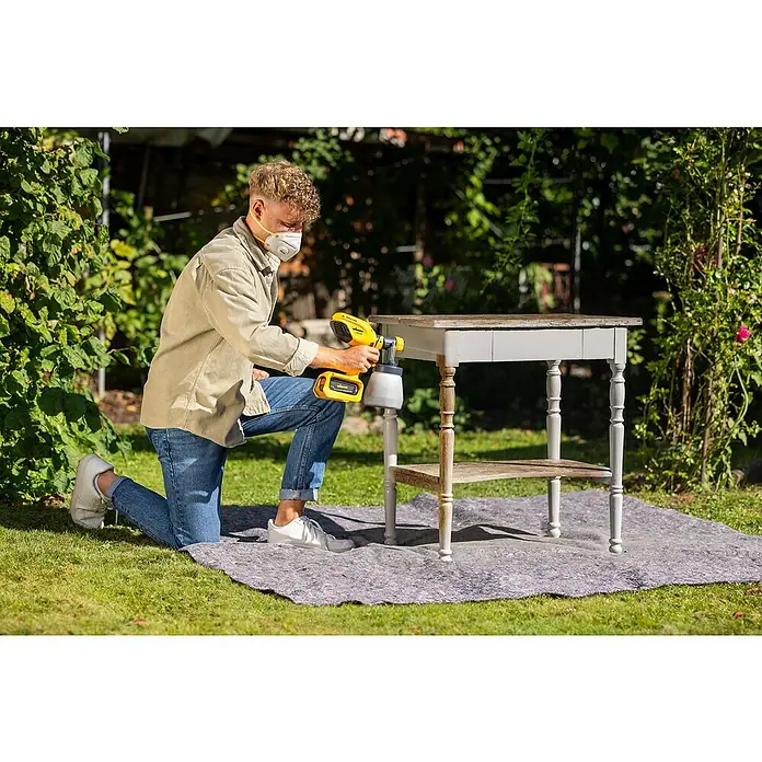 Wagner Akku-Farbsprühsystem Wood&Metal Sprayer W 250 18V - 18 V, Ohne Akku - Ein Mann kniet auf einer Decke und sprüht mit einer gelben Farbspritzpistole ein graues Beistelltischchen im Garten.