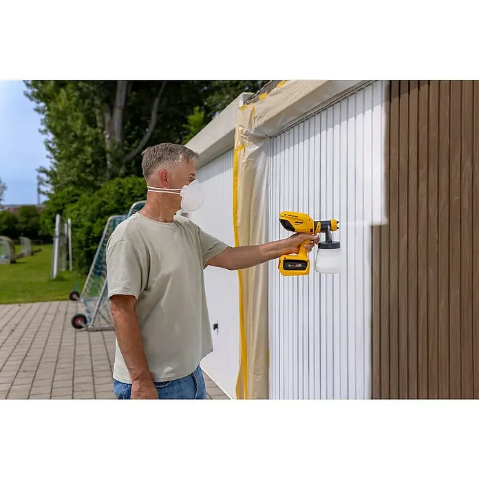 Wagner Akku-Farbsprühsystem Wood&Metal Sprayer W 250 18V - 18 V, Ohne Akku - Mann mit Atemschutzmaske lackiert eine Garagenwand mit einem gelben Wagner Farbsprühgerät. Das Ergebnis ist eine einheitliche, weiße Oberfläche.