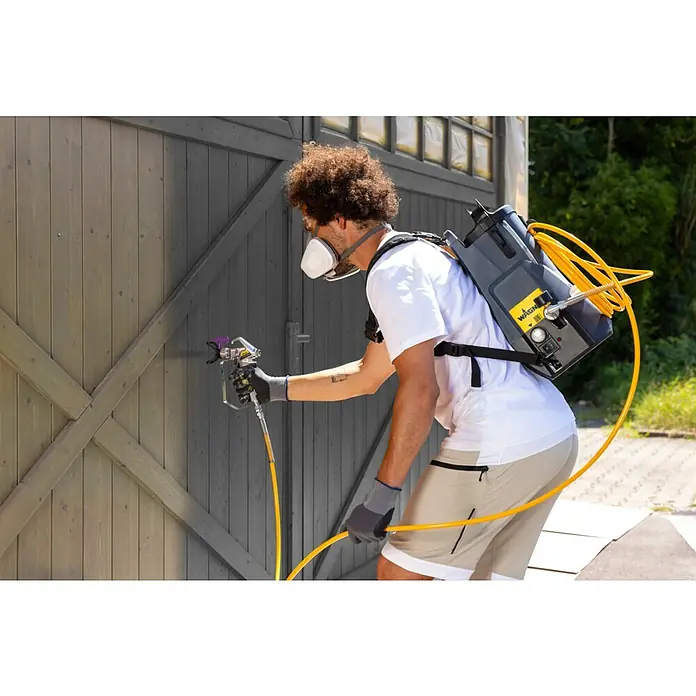 Akku-Farbsprühsystem - 18 V, 1 Akku, 4 Ah, Max. Fördermenge: 0,9 l/min - Ein Mann verwendet eine Wagner Airless Farbspritzpistole mit Rucksack, um ein Holztor zu streichen. Praktisch und effizient für große Flächen.