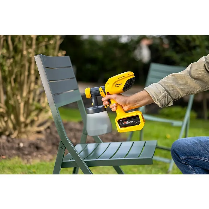 Wagner Akku-Farbsprühsystem Wood&Metal Sprayer W 250 18V - 18 V, Ohne Akku - Nahaufnahme einer Person, die einen gelben Wagner W 250 SE Akku-Farbspritzer in einem Garten benutzt, um einen grünen Stuhl zu bemalen.