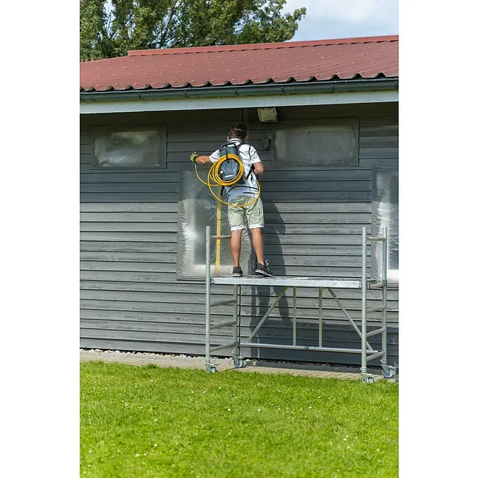 Akku-Farbsprühsystem - 18 V, 1 Akku, 4 Ah, Max. Fördermenge: 0,9 l/min - Ein Mann auf einem Gerüst sprüht Farbe auf die Fassade eines Hauses, während er einen Farbsprühgerät-Rucksack trägt.