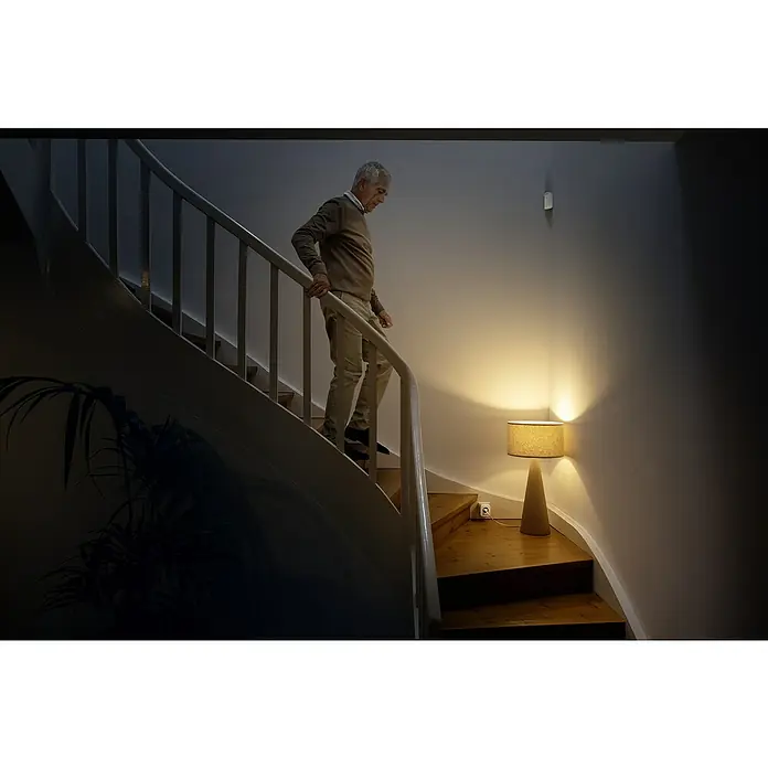Bosch Smart Home Bewegungsmelder - Weiß, Reichweite ca.: 50 m - Ein Mann geht eine Treppe hinunter, die von einer eleganten Lampe beleuchtet wird, die die Wand akzentuiert. Fokus liegt auf der Lampe.