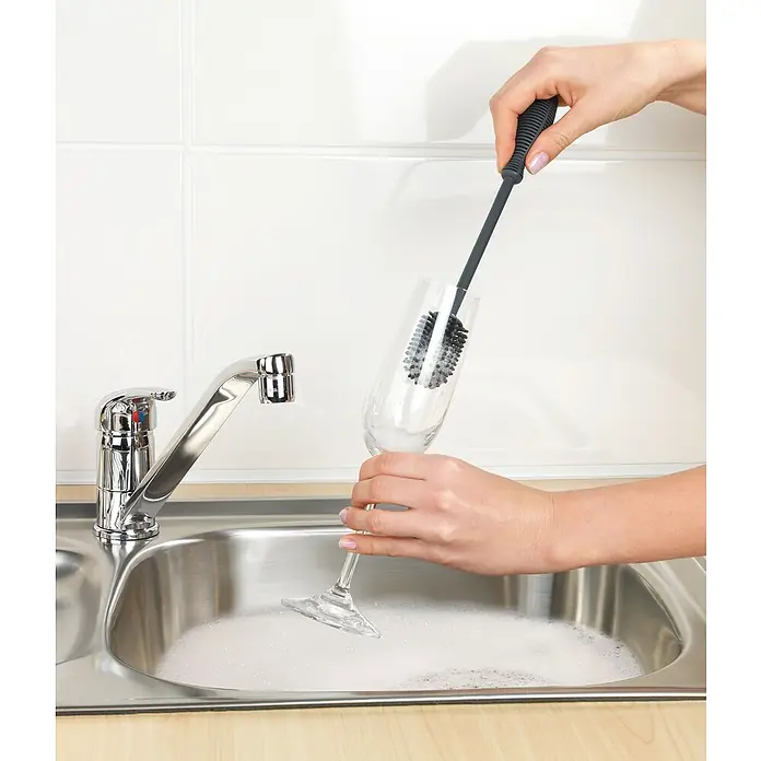 Wenko Spülbürste - Länge: 25 cm, Silikon - Eine Hand hält eine Flaschenbürste und reinigt ein Weinglas in einem mit Seifenwasser gefüllten Spülbecken vor einem weißen Fliesenspiegel.