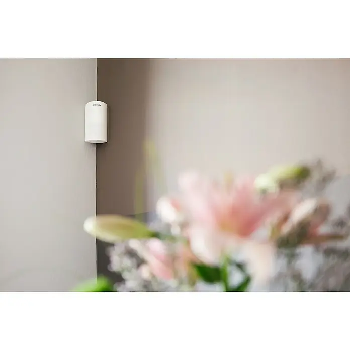 Bosch Smart Home Bewegungsmelder - Weiß, Reichweite ca.: 50 m - Ein weißer Bosch Bewegungsmelder ist an der Wand montiert und bietet zuverlässige Sicherheit im Raum. Die Blumen im Vordergrund sind unscharf.