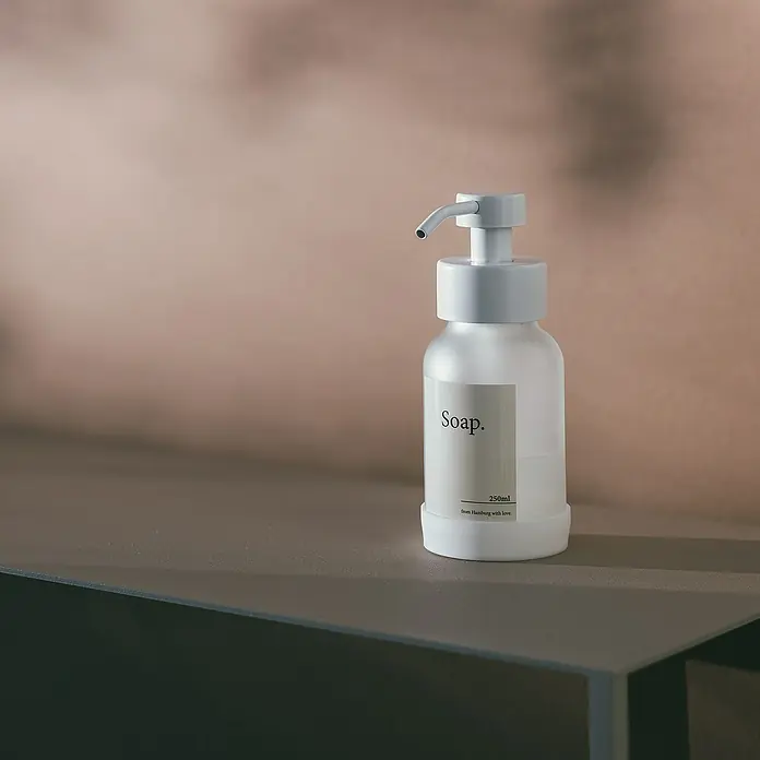 Designfabrik Hamburg Seifenspender - Edelstahl, Weiß/Satiniertes Glas - Eine minimalistische Seifenspenderflasche mit einem schlichten Etikett auf einem stilvollen Regal.