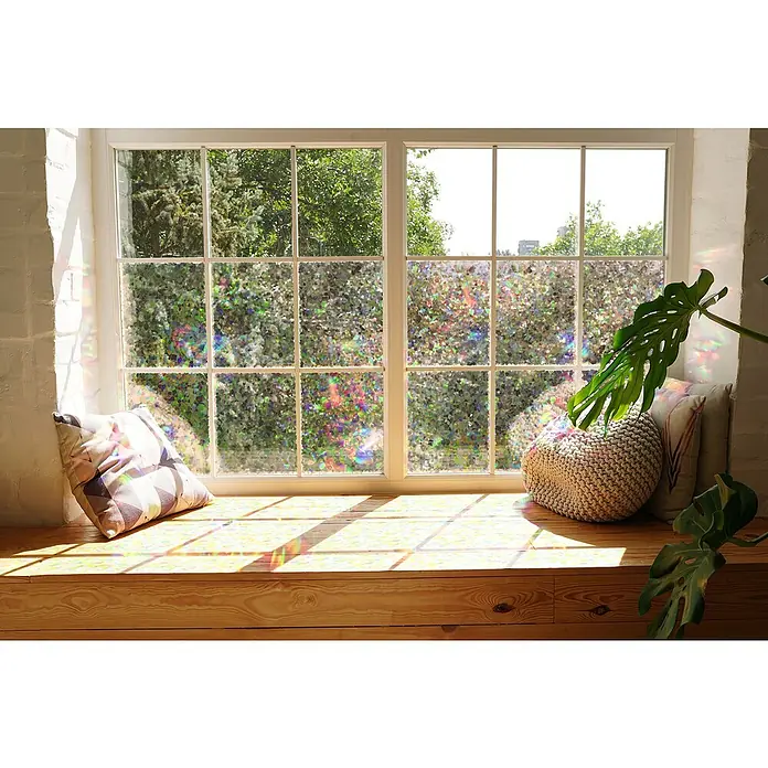 D-c-fix Static Glasfolie Rainbow Shards - 150 x 45 cm, Transluzent, Statisch haftend - Fensterbank mit Kissen und Pflanze vor einem Fenster mit reflektierender Fensterfolie, die das Sonnenlicht in Regenbogenfarben bricht.