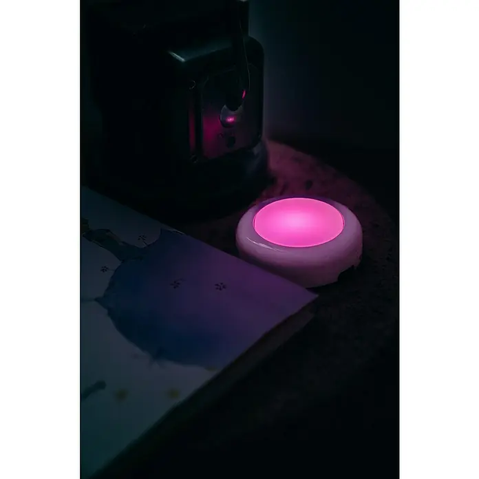 Osram Luz de armario LED Dot-It Twist & Shake - 1 luz, 0,5 W, Blanco - Un primer plano de una mesita de noche con una página de libro, una luz rosa redonda y un objeto negro al fondo.
