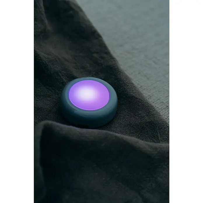 Osram Luz de armario LED Dot-It Twist & Shake - 1 luz, 0,5 W, Blanco - Primer plano de una luz nocturna redonda azul con un resplandor púrpura sobre un tejido gris.