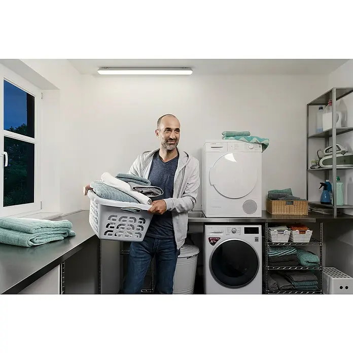Osram Tira de luz LED antihumedad Essential - Largo: 65,1 cm, IP65 - Hombre con cesto de ropa en la lavandería, con lavadoras Bosch al fondo. Diseño luminoso y moderno con amplio espacio de almacenamiento y estanterías.