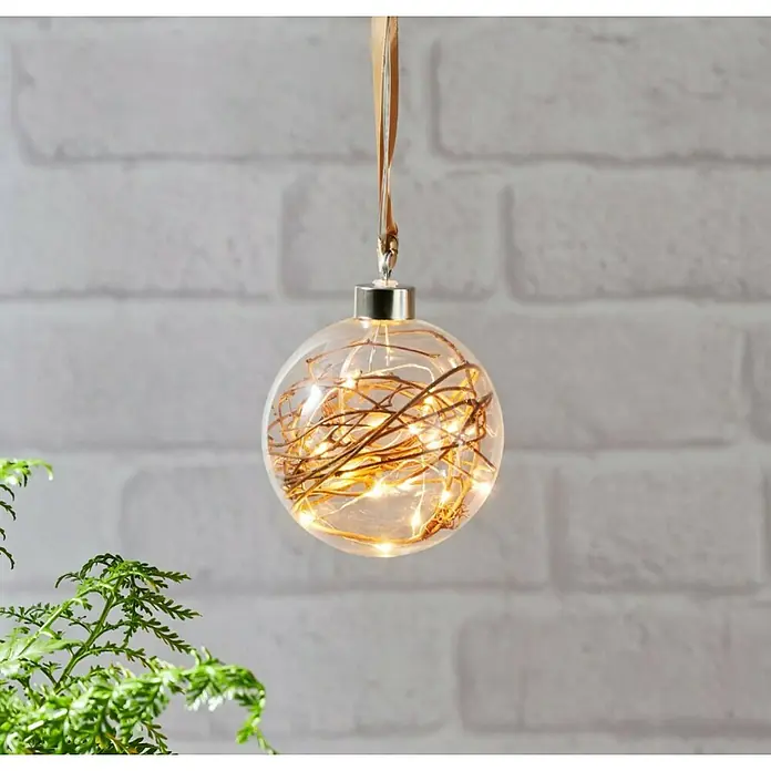 Eglo Lámpara colgante LED redonda Glow - 0,18 W, Diámetro: 10 cm, Blanco cálido, Dorado - Un adorno navideño transparente lleno de luces blancas cálidas y ramitas, colgado de una cinta contra una pared de ladrillos.