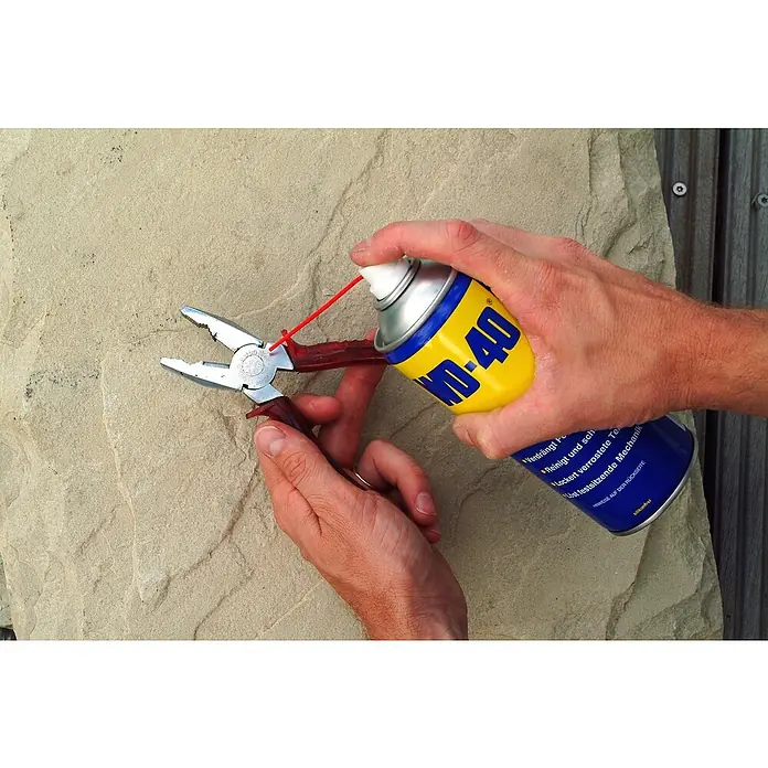 WD-40 Multiöl Classic - 100 ml - Sprühen von WD-40 auf eine Zange mit rotem Griff vor einer texturierten, beigen Oberfläche.
