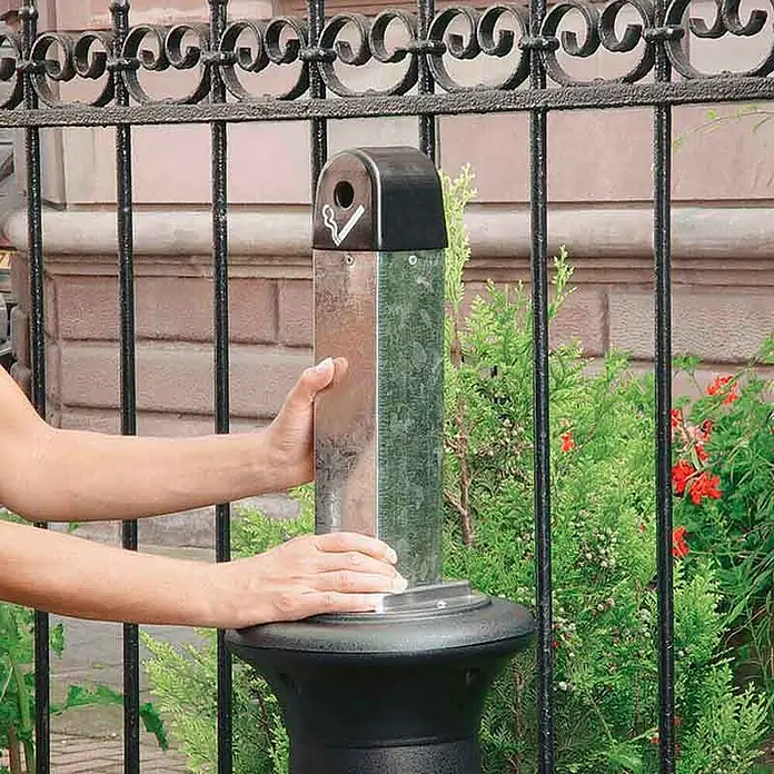 Proregal Asche-Mülleimer Rubbermaid Standaschenbecher mit Stahl-Inneneimer - Kunststoff, Schwarz - Nahaufnahme einer Person, die eine Metallsäule mit einem Aschenbecher berührt, eingebettet in ein dekoratives Eisengeländer.