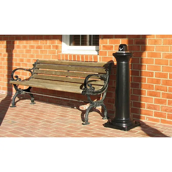 Proregal Asche-Mülleimer Rubbermaid Standaschenbecher mit Stahl-Inneneimer - Kunststoff, Schwarz - Eine Parkbank aus Holz und Metall steht neben einer schwarzen Säule vor einer roten Backsteinmauer. Die Bank lädt zum Verweilen ein.