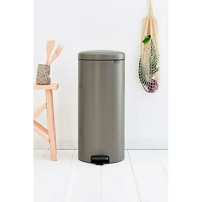 Proregal Treteimer Brabantia Klassischer Tretmülleimer mit Soft-Verschluss - 30 l, Metall, Platin - Ein stilvoller, grauer Treteimer von Brabantia steht auf einem weißen Holzboden, neben einem Hocker und einer Einkaufstasche.