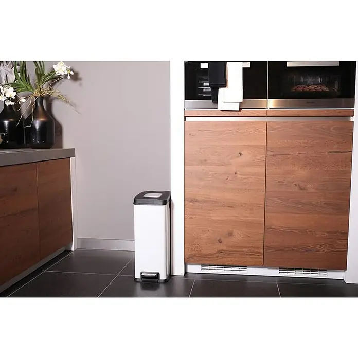 Proregal Treteimer Schmaler rechteckiger Pedaleimer mit Softclose-Deckel - 30 l, Kunststoff, Weiß - Ein moderner, weißer Treteimer steht neben einer Küchentür aus Holz mit zwei eingebauten Öfen darüber. Der Treteimer ist schlank und rechteckig.