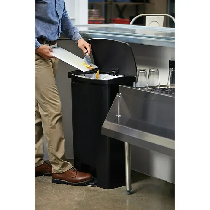Proregal Treteimer Rubbermaid Slim Jim Step On Container Front Step Pedal-Abfalleimer - 68 l, Kunststoff, Schwarz - Bild eines schwarzen Treteimer, der von einer Person benutzt wird, die Abfälle entsorgt. Der Fokus liegt auf dem funktionalen Design des Eimers.