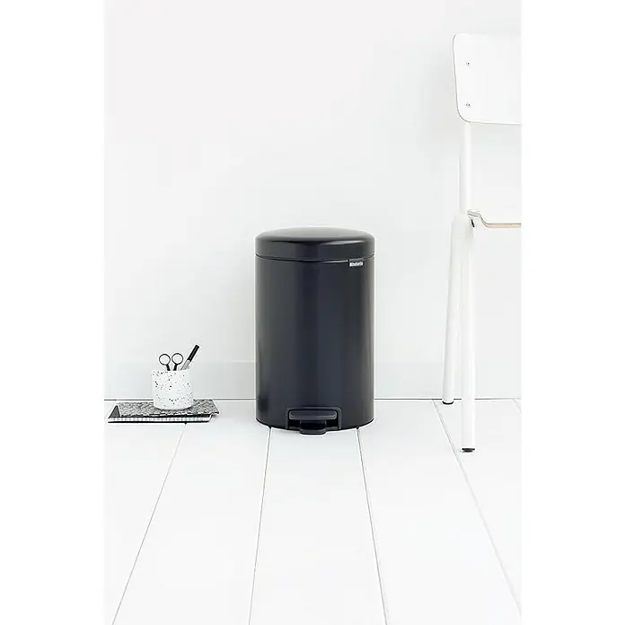 Proregal Treteimer Brabantia Klassischer Tretmülleimer mit Soft-Verschluss - 12 l, Metall, Metallik Schwarz - Diese Nahaufnahme zeigt einen stilvollen, schwarzen Treteimer von Brabantia vor einer minimalistischen Wand und einem weißen Stuhl.