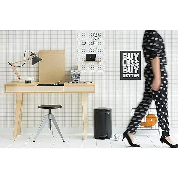 Proregal Treteimer Brabantia Klassischer Tretmülleimer mit Soft-Verschluss - 12 l, Metall, Metallik Schwarz - Schreibtisch mit Lampe, Stuhl und Papierkorb vor einer Wand mit Gittermuster, daneben eine Person in gepunktetem Outfit.