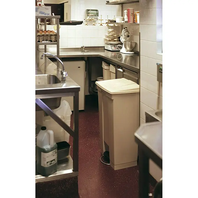 Proregal Treteimer Rubbermaid Robuster Pedaleimer - 87 l, Kunststoff, Beige - Ein beiger Mülleimer mit Deckel steht in einer gewerblichen Küche mit rotem Boden und Edelstahlarbeitsflächen.