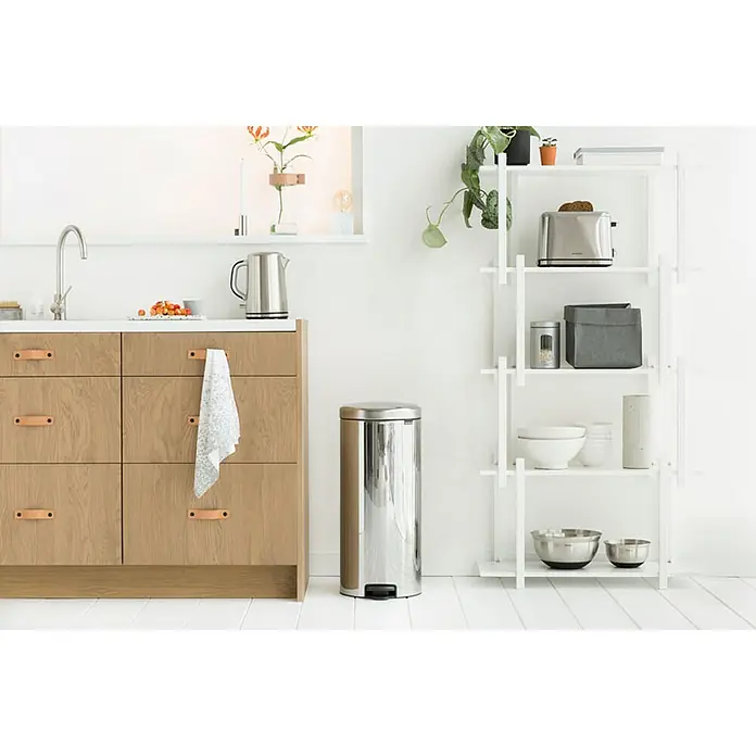 Proregal Treteimer Brabantia Klassischer Tretmülleimer mit Soft-Verschluss - 30 l, Metall, Edelstahl - Küchenszene mit einem Edelstahl-Treteimer im Vordergrund, einer Kommode mit hellen Holzfronten und einem weißen Regal mit Küchenutensilien.