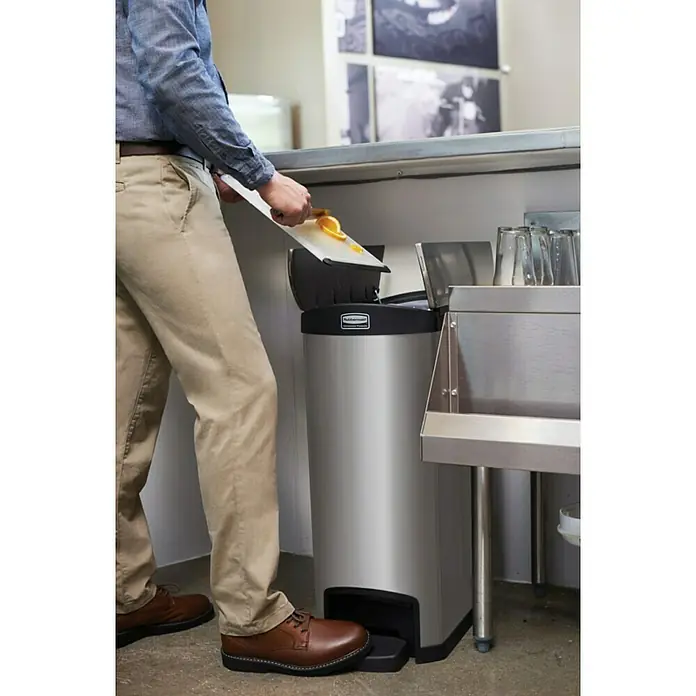 Proregal Treteimer Rubbermaid Slim Jim Step On Container End Step Pedal-Abfalleimer - 50 l, Kunststoff, Silber mit schwarzem Akzentstreifen - Eine Person wirft Abfälle in einen silbernen Rubbermaid-Treteimer mit Fußpedal. Der Abfalleimer steht in einer gewerblichen Küche.
