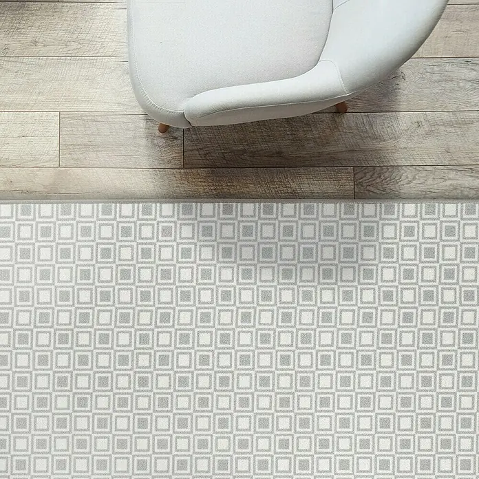 Teppichläufer Bray - Grau, 150 x 80 cm, 100 % Polyamid - Ein grauer Teppich mit einem quadratischen Muster liegt auf dem Boden neben einem Sessel. Der Teppich hat ein modernes und stilvolles Design.