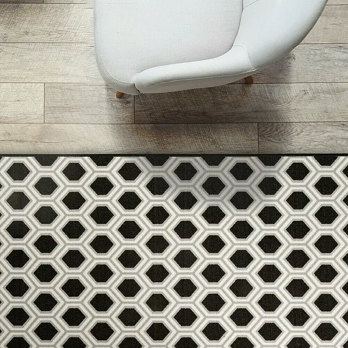 Teppichläufer Bangor - Schwarz/Weiß, 350 x 80 cm, 100 % Polyamid - Blick von oben auf einen schwarz-grauen Teppich mit geometrischem Muster unter einem hellen Sessel auf einem Holzboden.