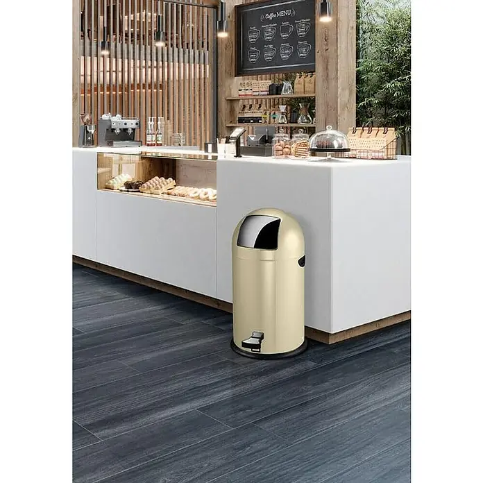 Proregal Treteimer - 33 l, Stahl, Creme - Ein beigefarbener, runder Treteimer aus Metall steht vor einer weißen Theke in einem Café oder Restaurant. Der Eimer ist sauber und modern.