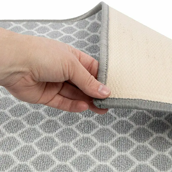 Teppichläufer Tralee - Grau, 300 x 80 cm, 100 % Polyamid - Detailansicht eines grauen Teppichs mit geometrischem Muster und einem beigen, rutschfesten Rücken, der von einer Hand angehoben wird.
