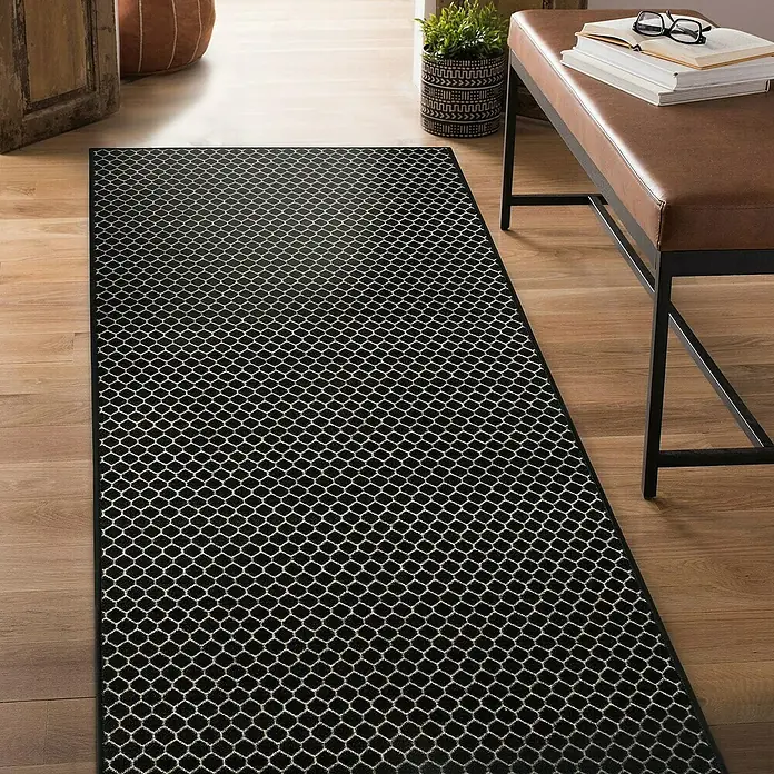 Teppichläufer Dundalk - Schwarz/Weiß, 150 x 80 cm, 100 % Polyamid - Ein schwarzer Teppich mit einem weißen Gittermuster auf einem Holzboden, neben einer braunen Bank und einer Topfpflanze.