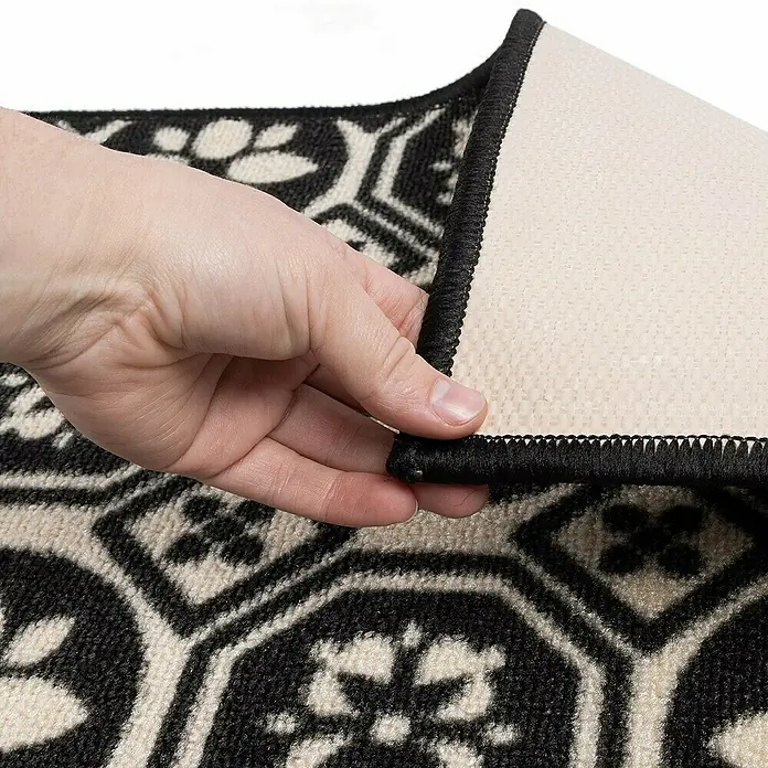 Teppichläufer Asaph - Schwarz/Weiß, 200 x 80 cm, 100 % Polyamid - Detailaufnahme eines schwarzen und beigen Teppichs mit geometrischem Muster, dessen Rückseite von einer Hand angehoben wird.