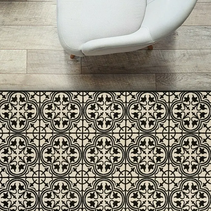 Teppichläufer Galway - Schwarz/Creme, 300 x 80 cm, 100 % Polyamid - Detailaufnahme eines dekorativen, schwarz-weißen Teppichs mit floralem Muster auf einem Holzboden, teilweise von einem Sessel bedeckt.