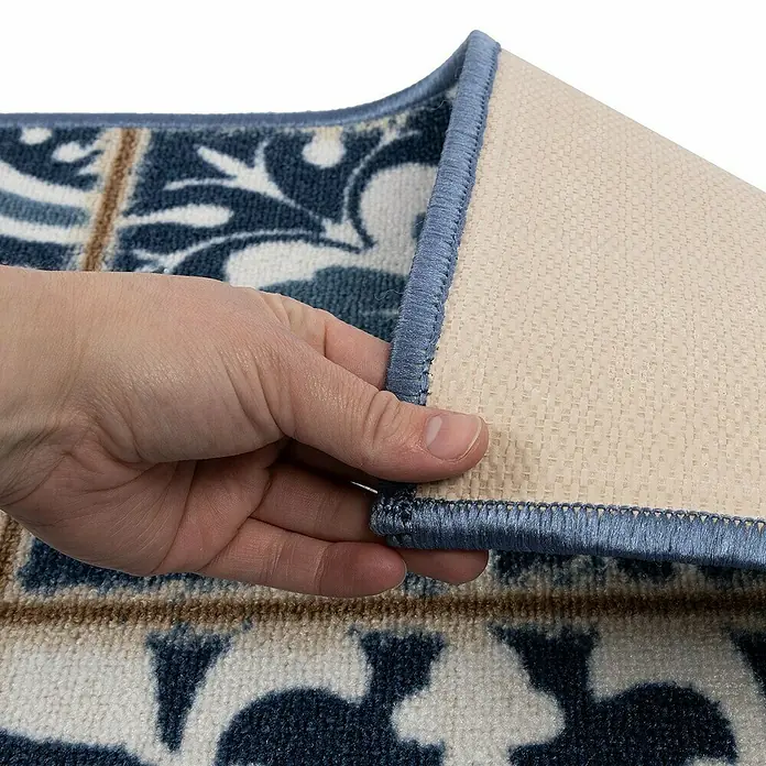 Teppichläufer Swansea - Blau, 250 x 80 cm, 100 % Polyamid - Nahaufnahme eines Teppichs mit blau-weißem Muster, der von einer Hand angehoben wird, um die Unterseite mit blauem Rand zu zeigen.